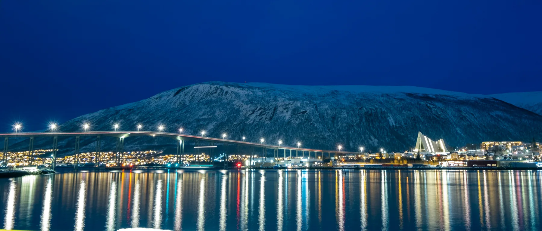 Osvětlený most v Tromsø a Arktická katedrála pod zasněženou horou, odrážející se v průlivu v modré hodině