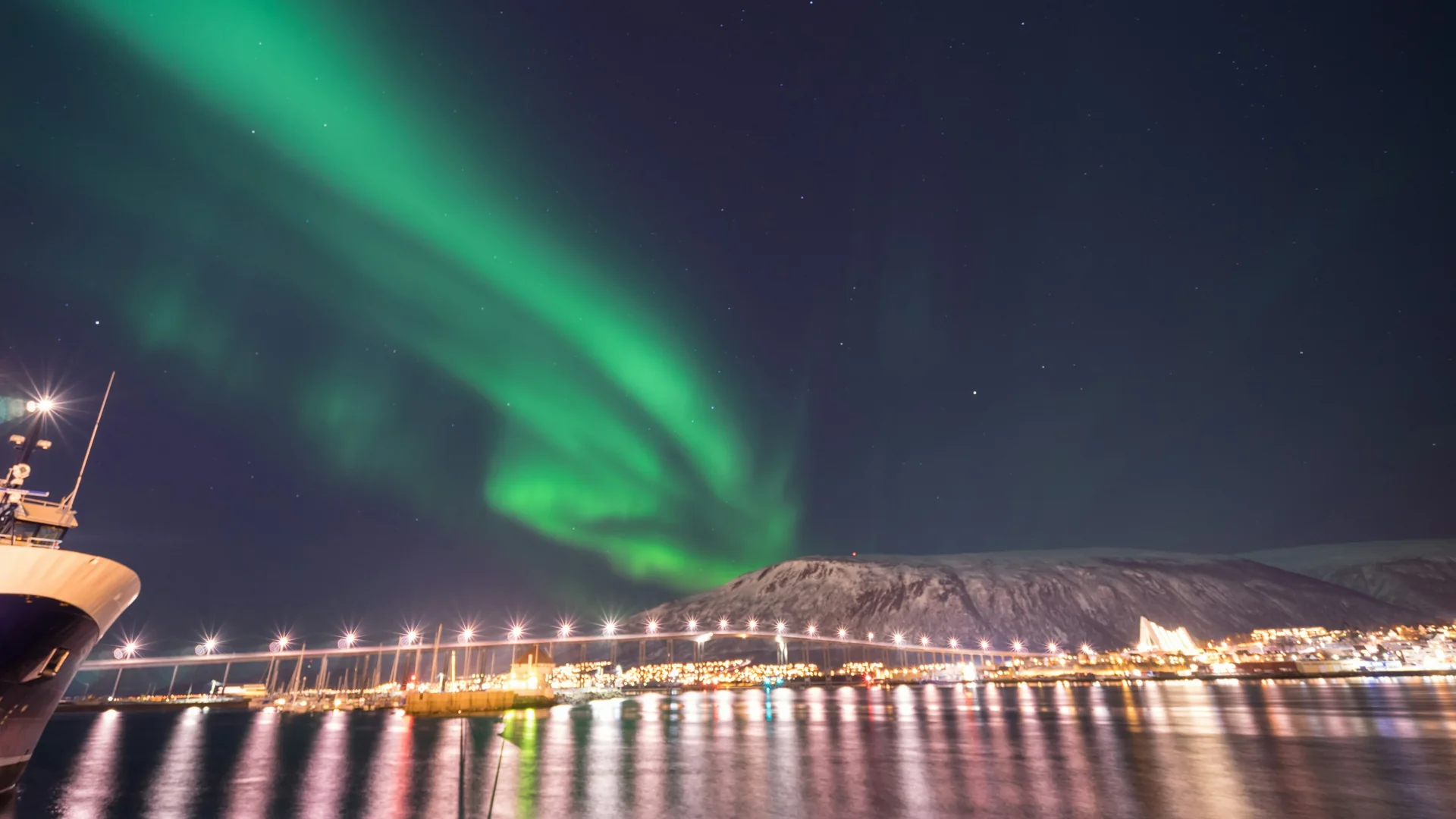 Zelená polární záře klenoucí se nad osvětleným mostem v Tromsø a Arktickou katedrálou, viděná v noci přes přístav